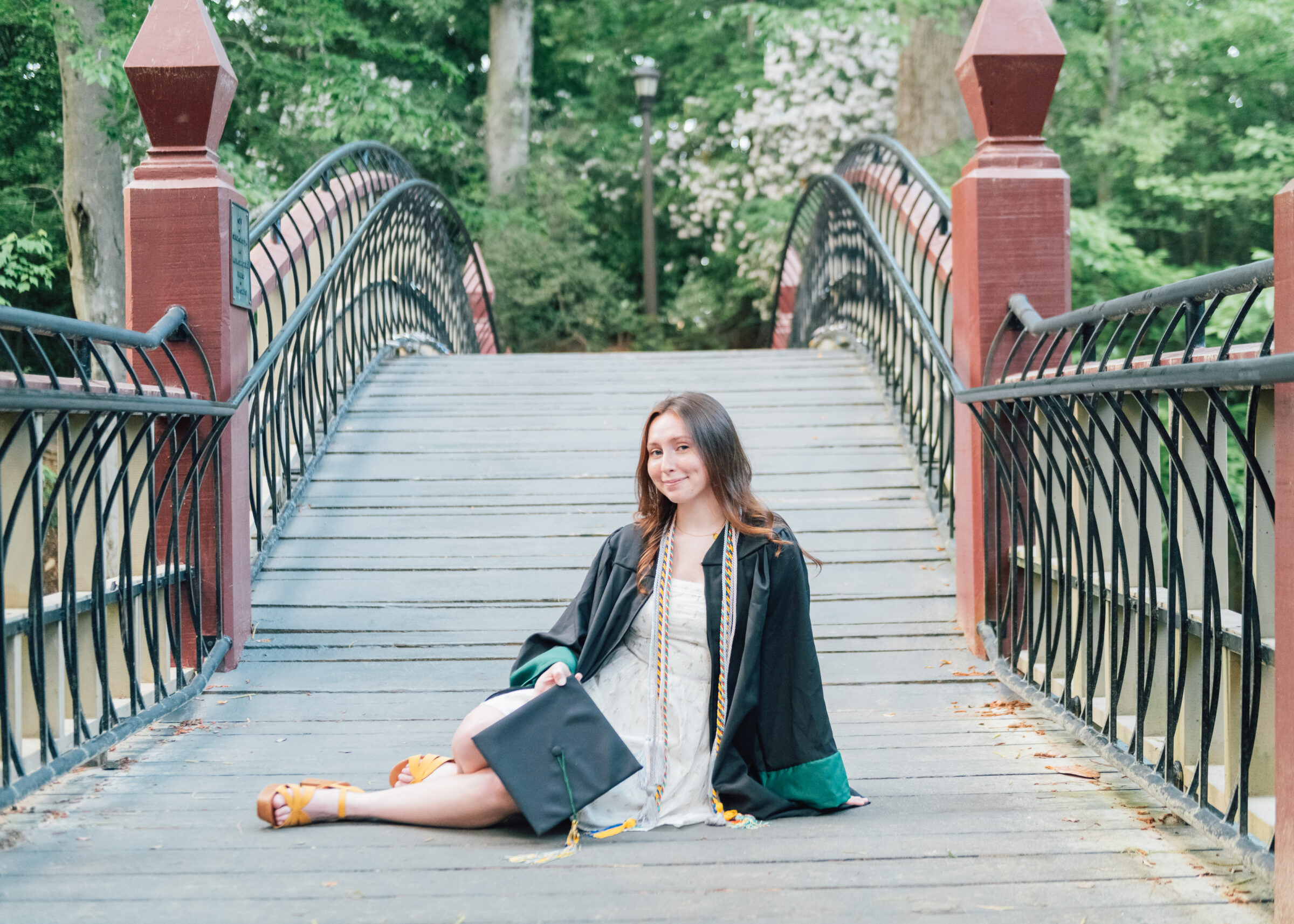 Graduate student in a black gown sits on a wooden bridge, cap in hand, with red rail posts and lush green trees in the background.