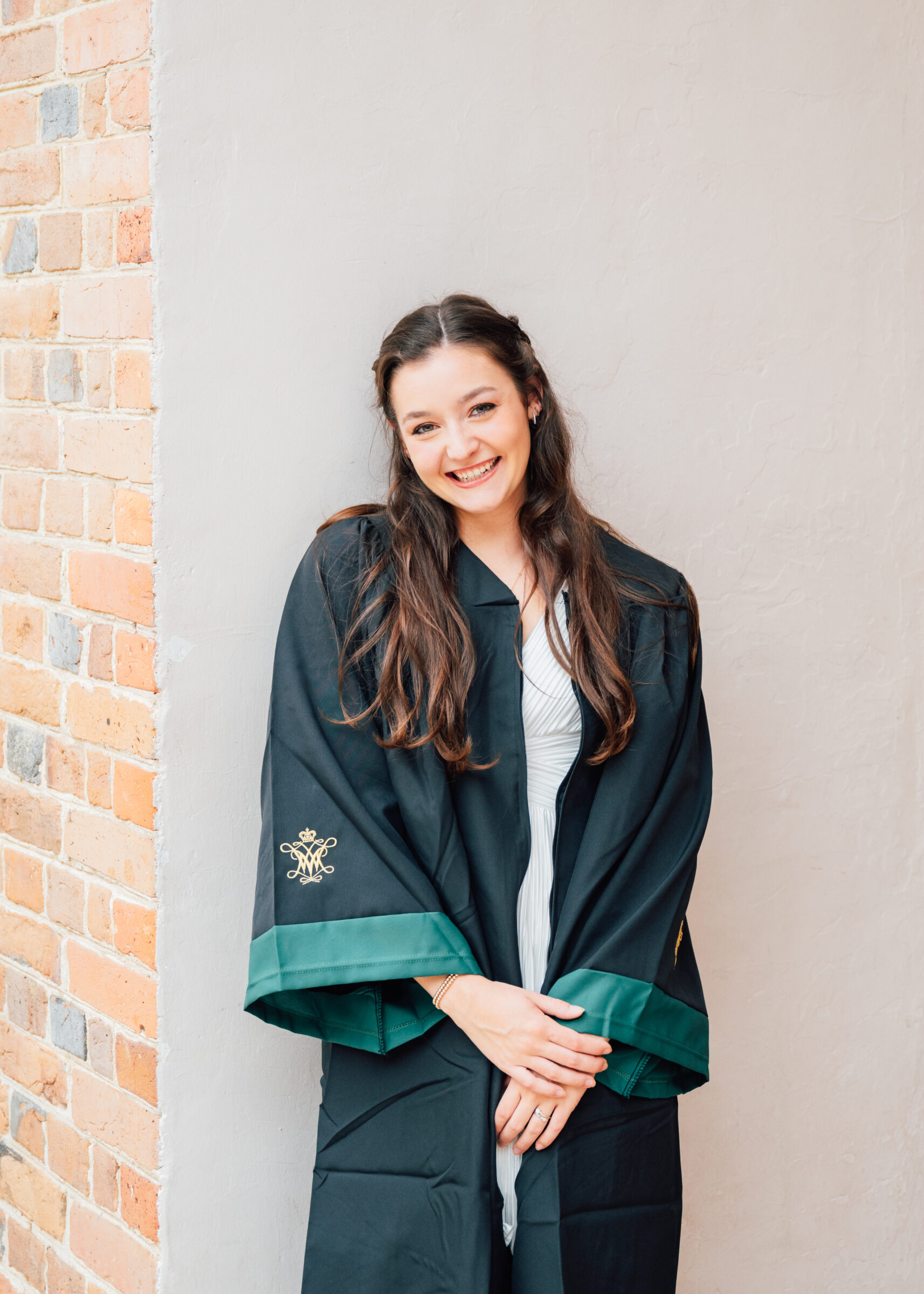 Smiling woman in a dark graduation gown leaning against a brick wall.