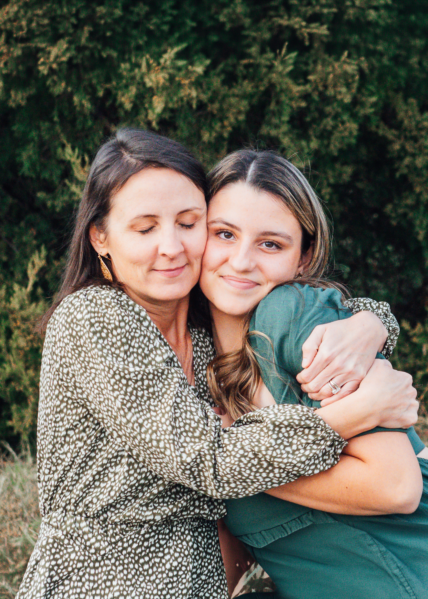 Two women embrace outdoors, one with eyes closed and a gentle smile, the other looking at the camera, green foliage behind them.